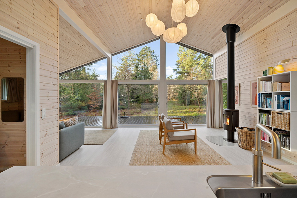 Stenhøj Husene designlinje Lux. Indvendig fra stue med pejs gennem store vinduer i hvid.