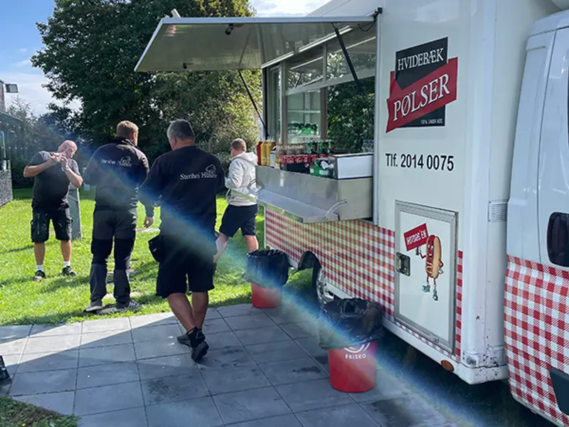 Solstråler henover forbi pølsevognen imens glad håndværkere spiser en hotdog.